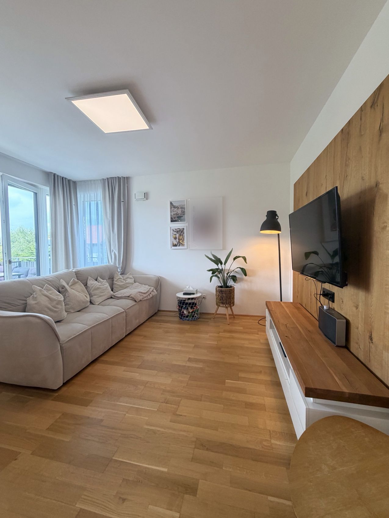 Wohnbereich mit heller Couch, Eichenwand mit Fernseher und großen Fenstern zum Balkon